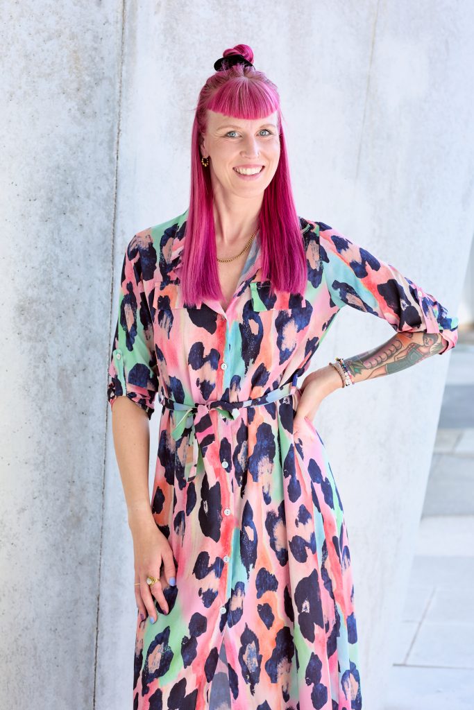 Frau, Sarah Blume mit rosa Haaren und buntem Kleid, freundliche lächelnd an einer Betonwand angelehnt.